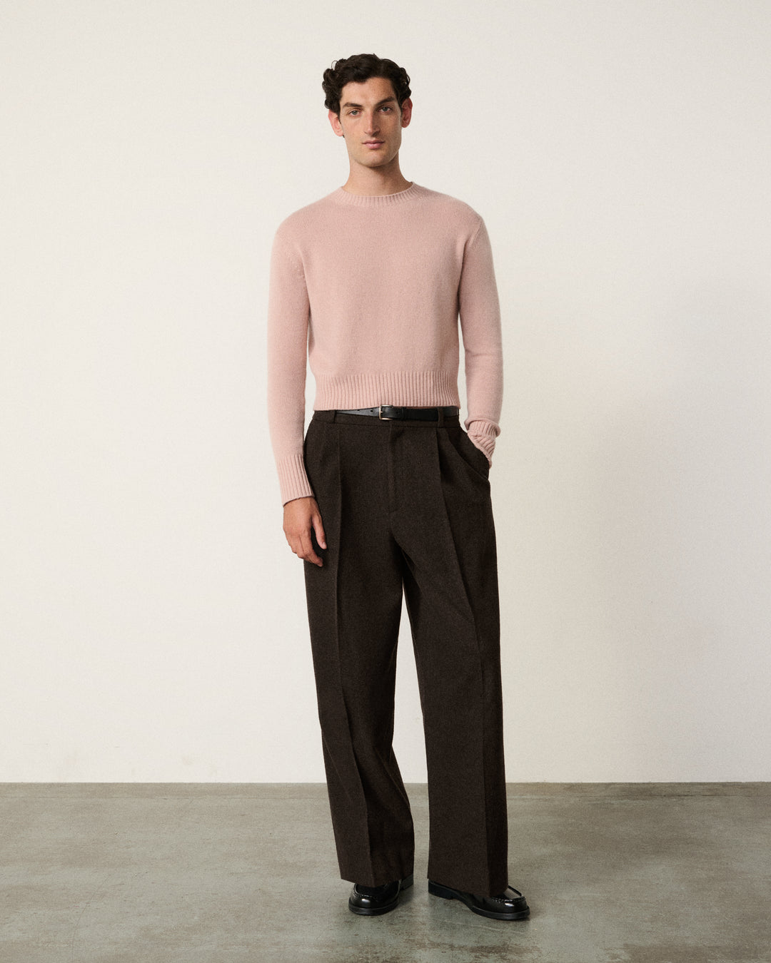 A man stands in a studio wearing a light pink long-sleeve knit sweater and dark brown wide-leg trousers, paired with a black belt and black shoes.