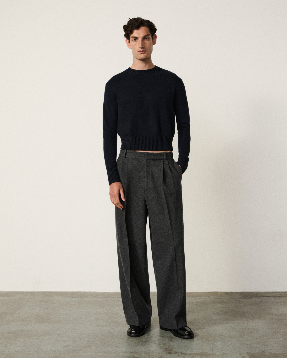 A male model stands in a studio wearing a black long-sleeve knit top and dark charcoal gray wide-leg trousers, paired with black shoes, against a plain off-white wall and concrete floor.