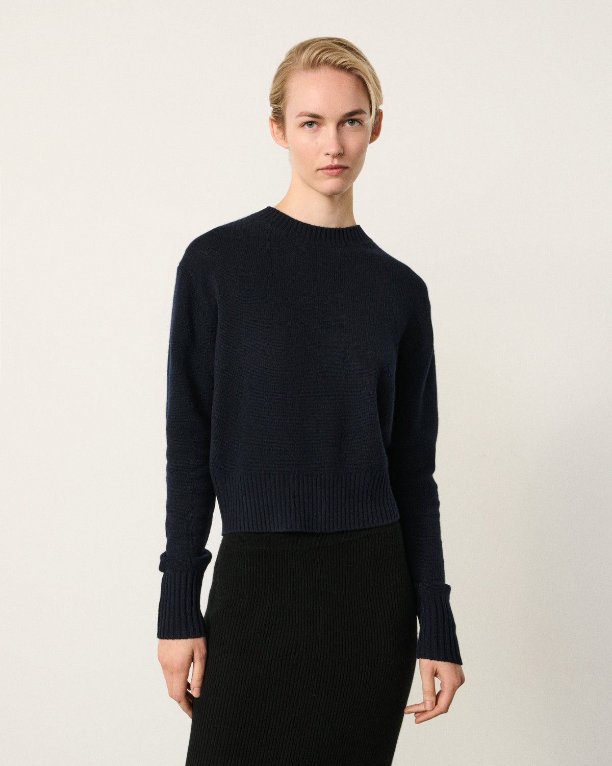 A model wearing a dark navy blue long-sleeve knit sweater with a crew neck and ribbed hem/cuffs, paired with a black ribbed skirt, against a plain off-white studio background.