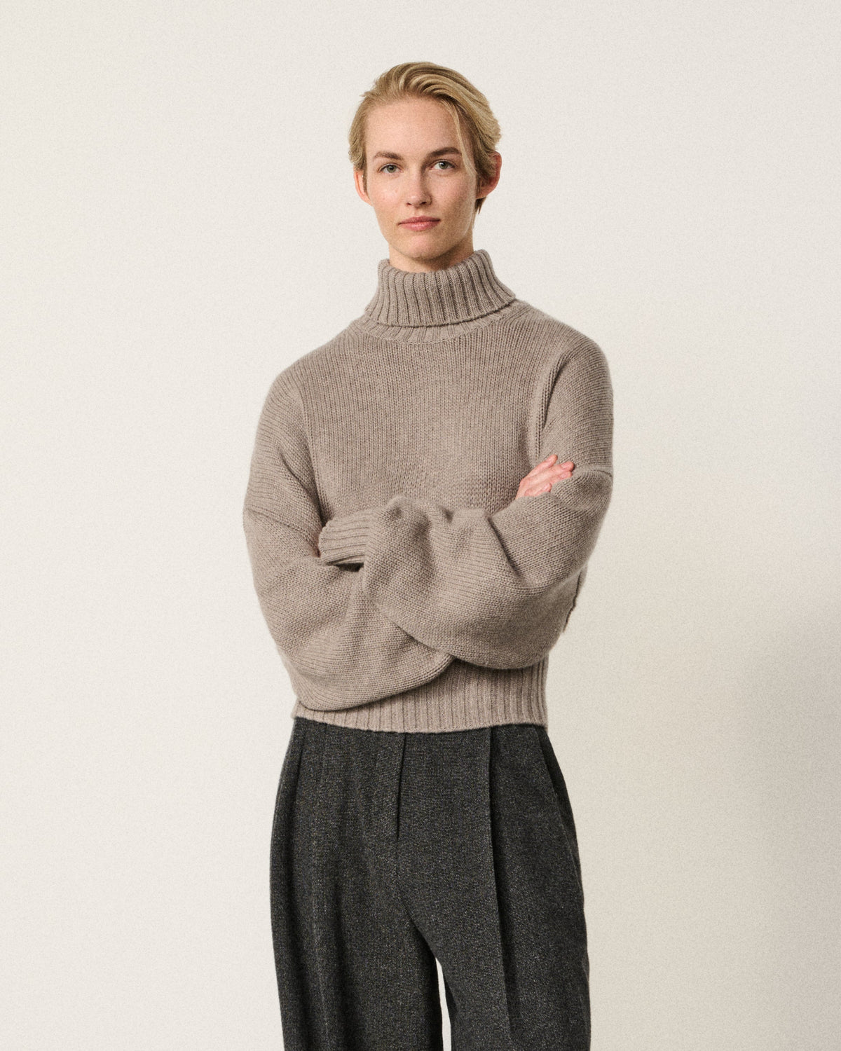 A woman stands with arms crossed wearing a light taupe knit turtleneck sweater and dark charcoal wide-leg trousers against a plain off-white studio wall and concrete floor.