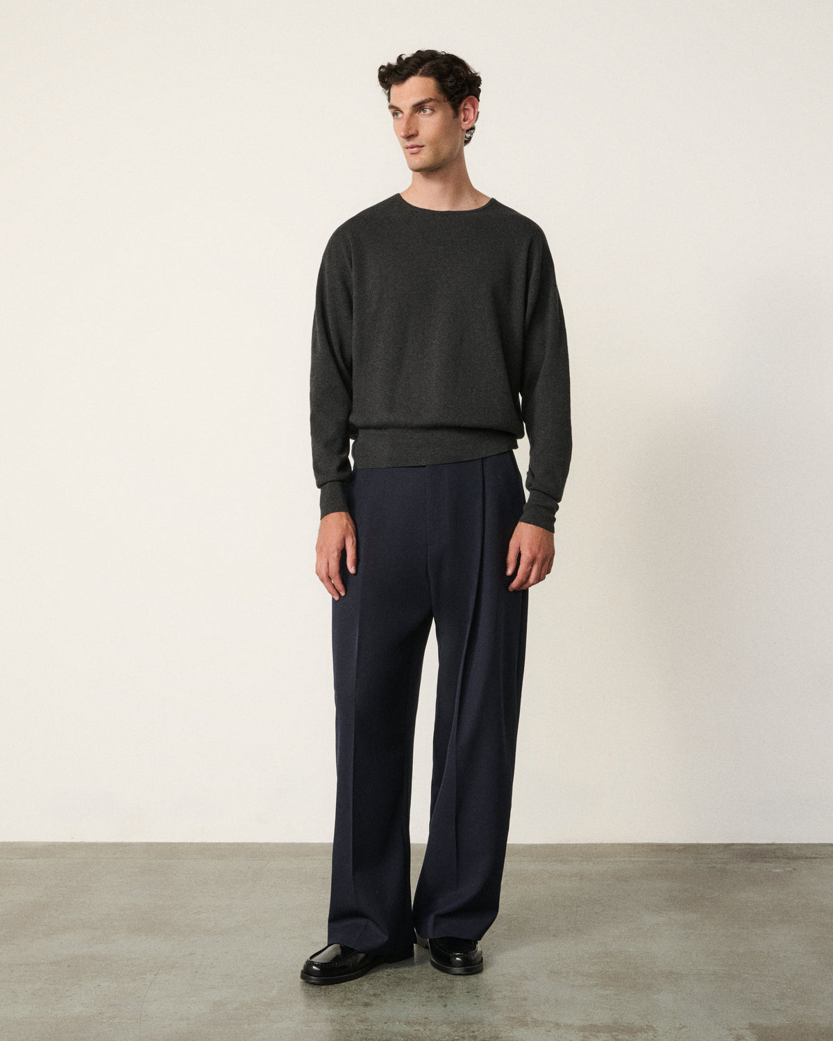 A man stands in a studio wearing a dark charcoal gray crewneck sweater and dark navy wide-leg trousers with black loafers.