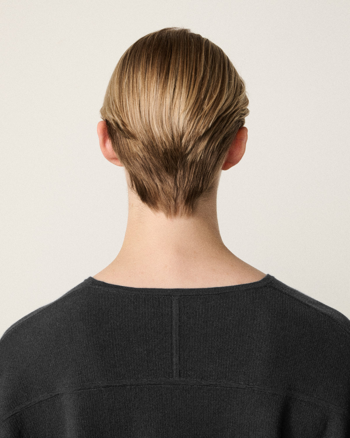 Back view of a person with styled light brown hair pulled back, wearing a dark charcoal gray top against a plain off-white background.