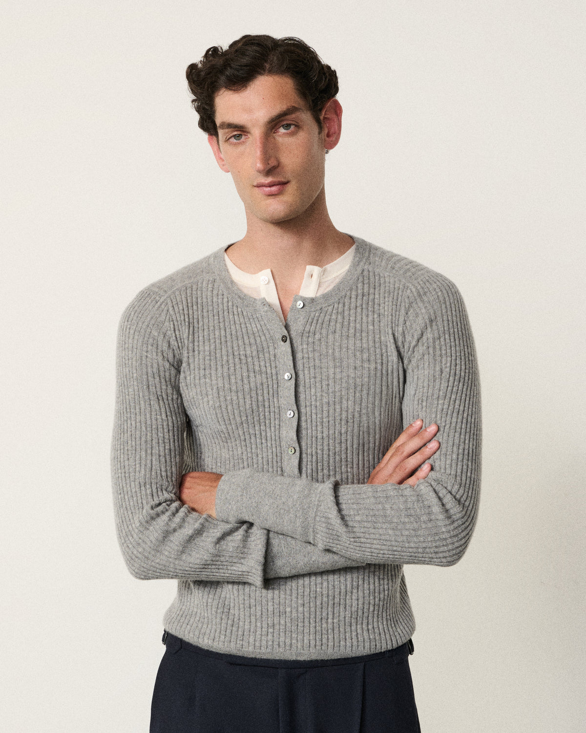 A man wearing a light gray ribbed knit cardigan over a white button-down shirt, paired with dark trousers, standing against a plain off-white background.