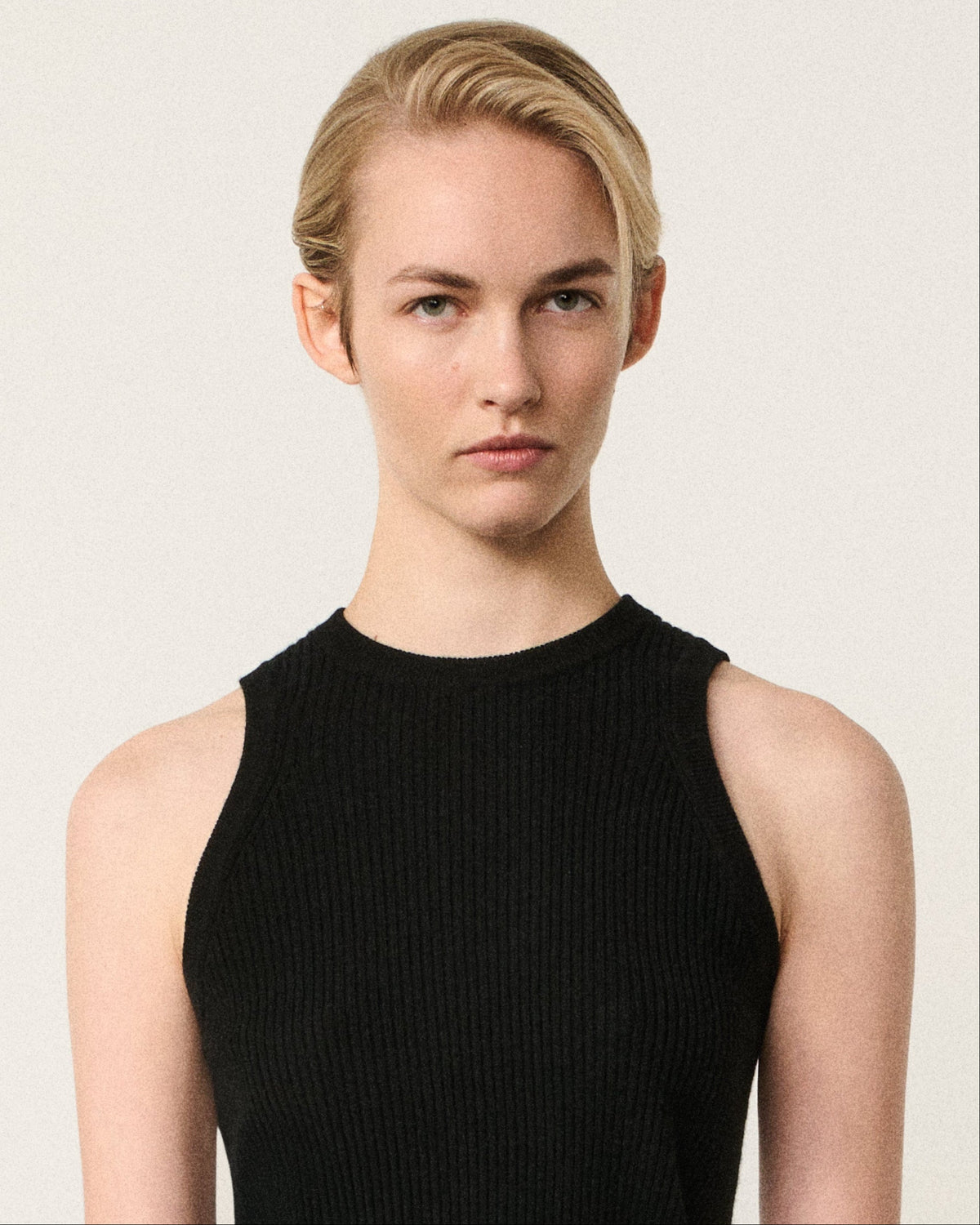 A model wearing a black ribbed sleeveless mock-neck top and high-waisted gray pleated wool trousers, photographed against a solid off-white studio background.