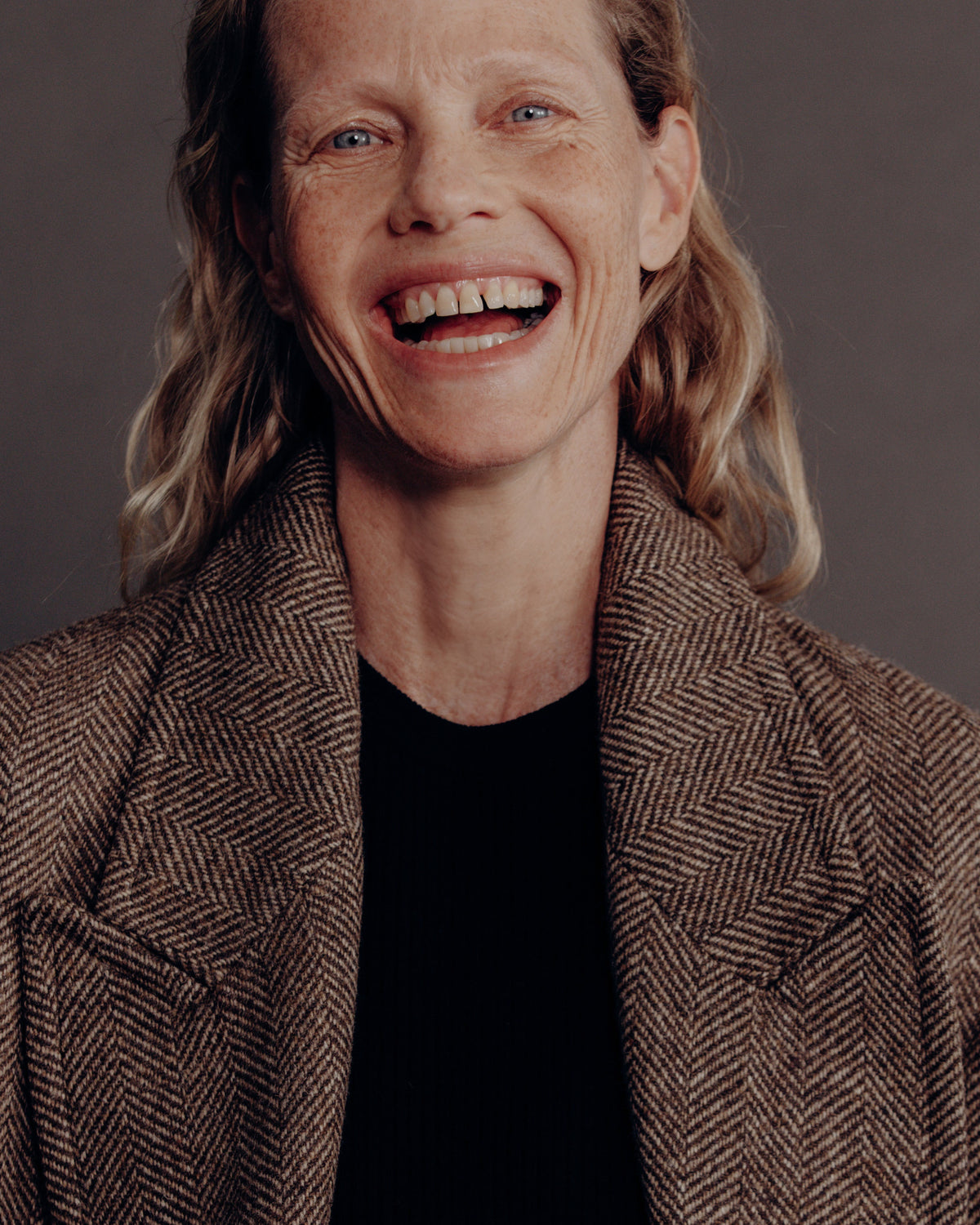 A smiling woman wearing a brown and beige herringbone patterned coat over a black top, photographed against a dark gray studio background.