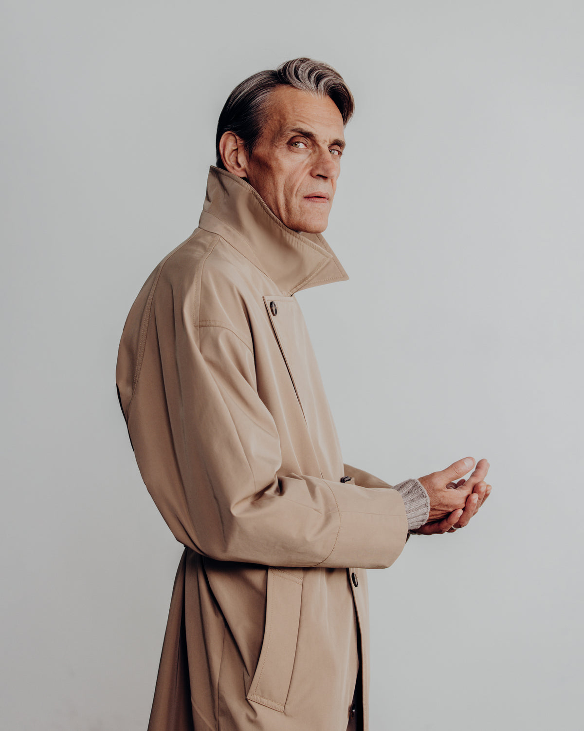 A man in profile wearing a beige trench coat over a gray ribbed sweater, photographed against a plain light gray studio background.