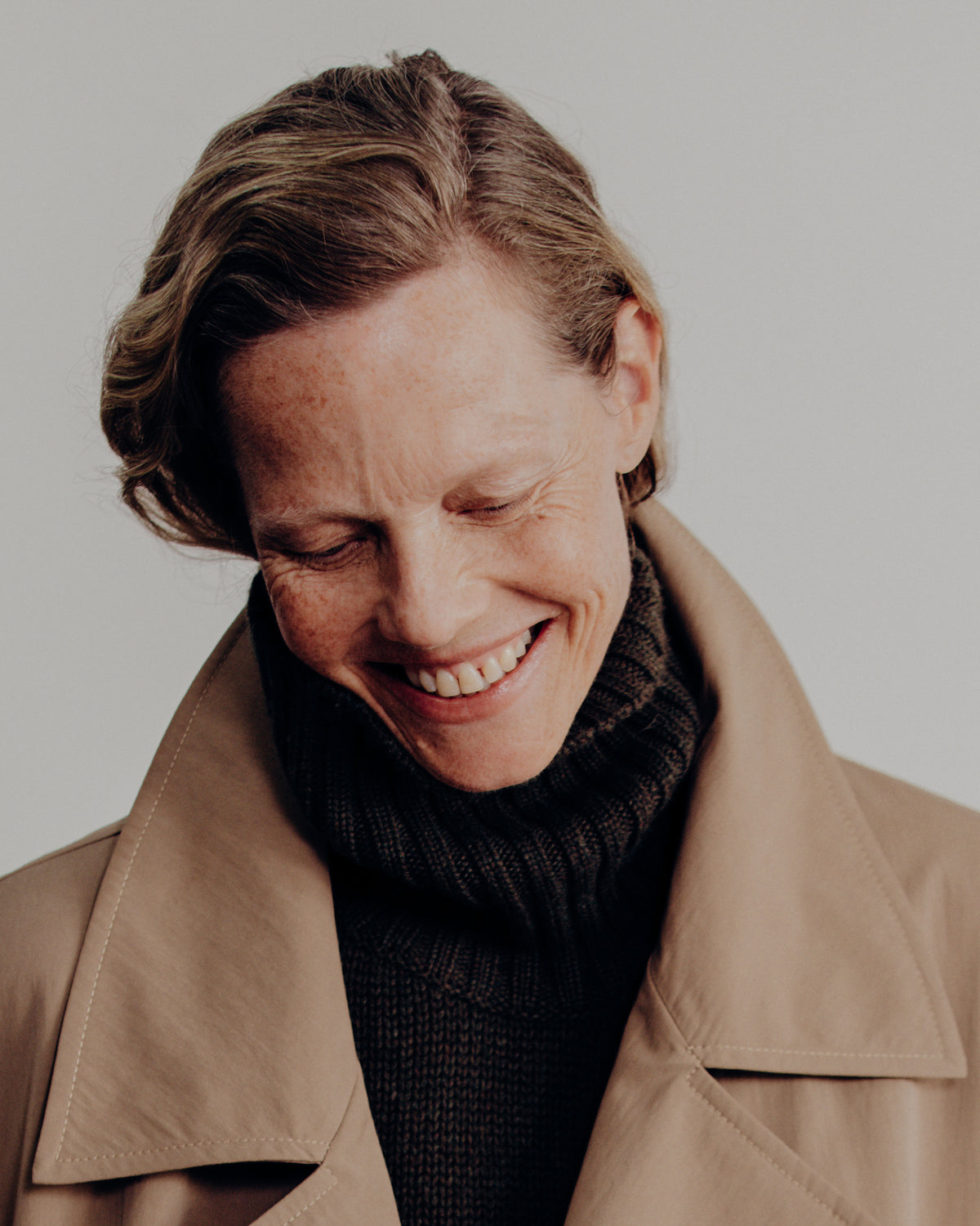 A close-up portrait of a smiling woman with short brown hair, wearing a beige trench coat over a black ribbed turtleneck sweater, set against a plain light gray background.