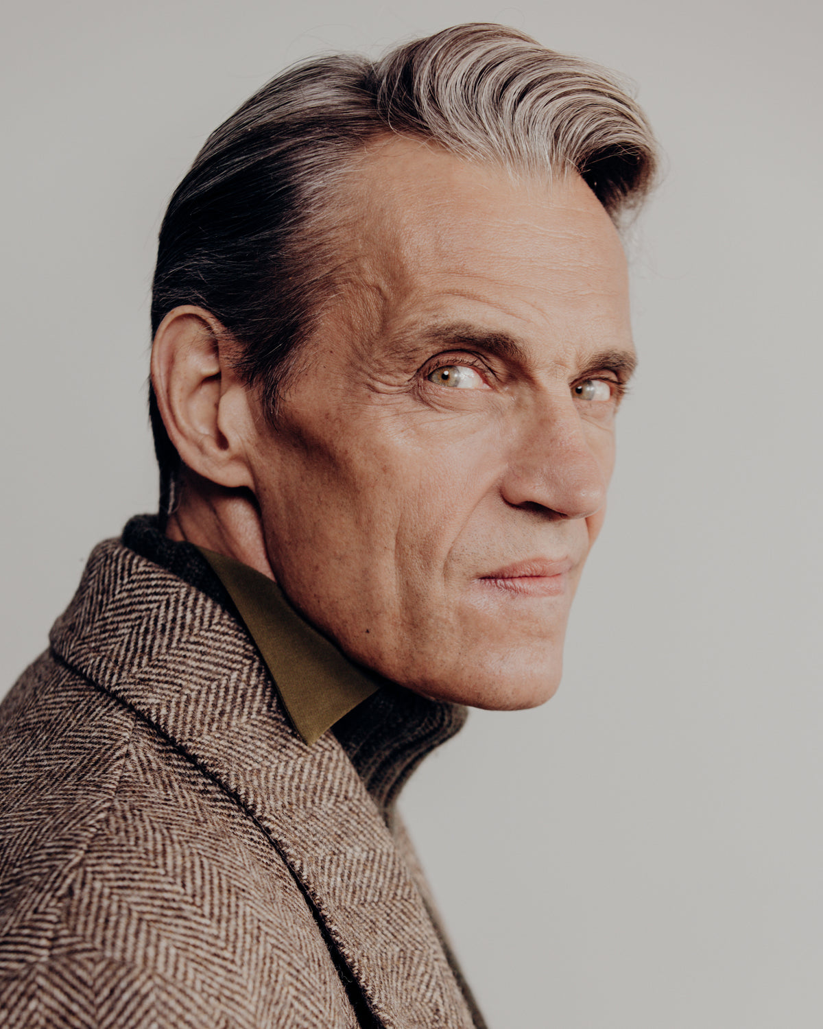 A close-up portrait of a man with gray hair wearing a brown herringbone patterned jacket over a dark olive shirt, set against a solid light gray background.