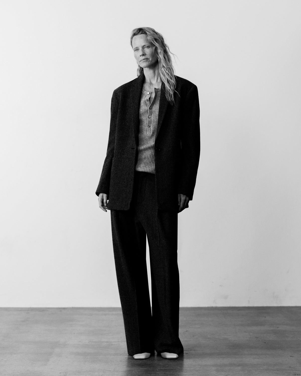 A woman stands in a minimalist studio wearing a dark oversized blazer, matching wide-leg trousers, a textured knit top, and pointed shoes against a plain light wall.