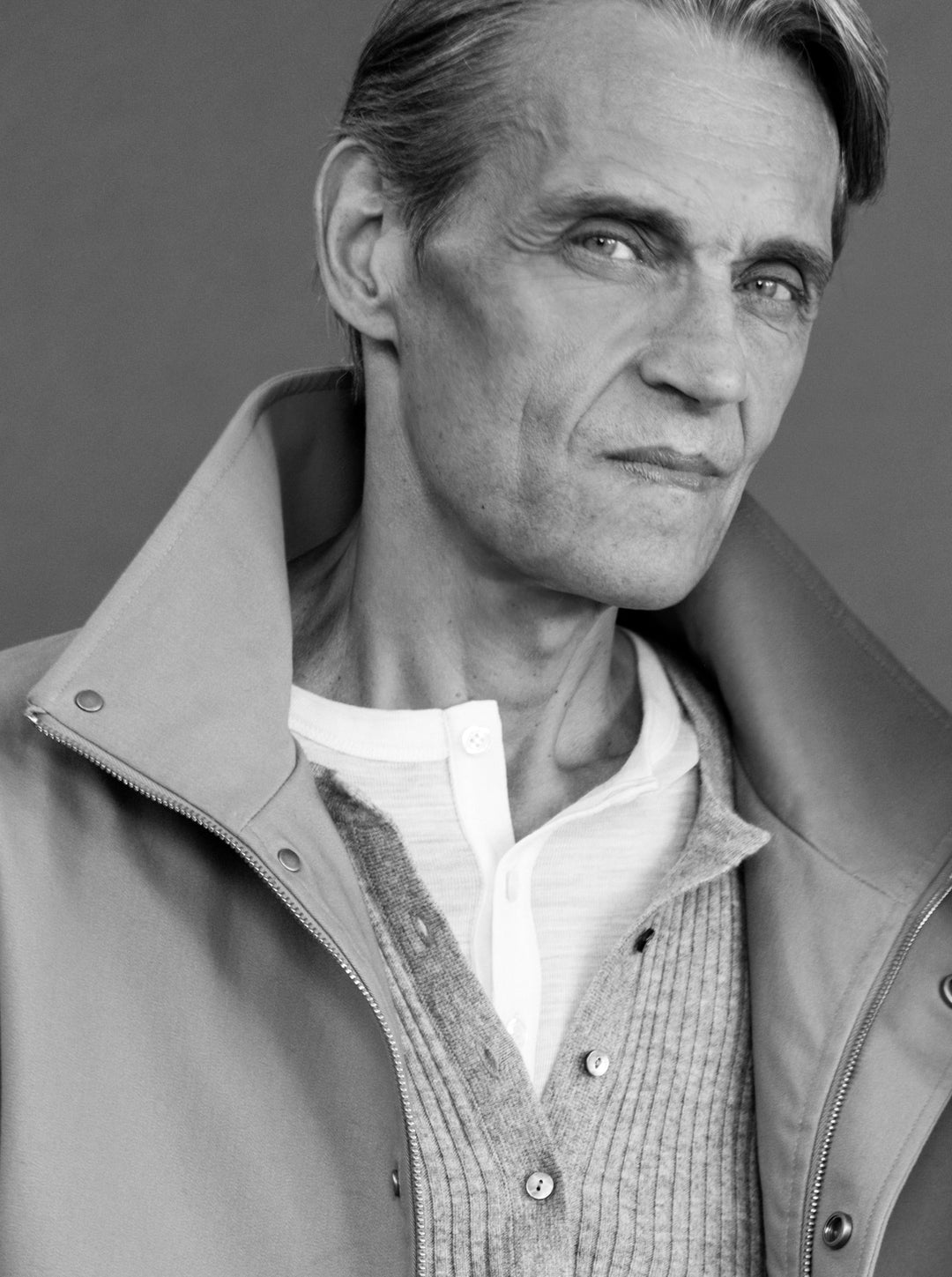 A close-up black and white portrait of a man wearing a light-colored zip-up jacket over a textured knit cardigan and white henley shirt, set against a neutral gray background.